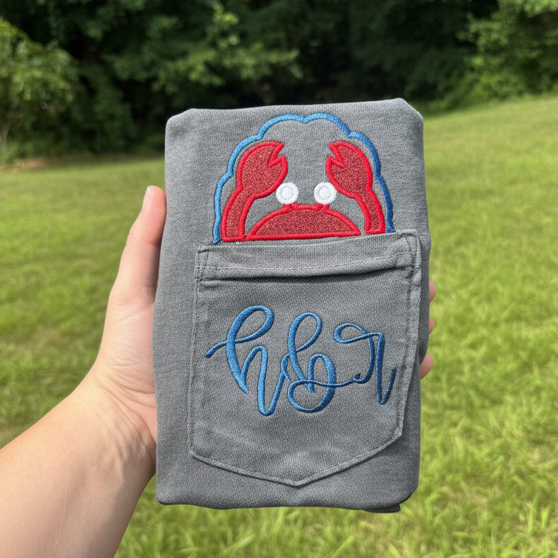Gray Comfort Colors pocket t-shirt featuring a red crab applique peeking above the pocket with a personalized embroidered monogram stitched across the pocket, coastal beach-themed apparel.