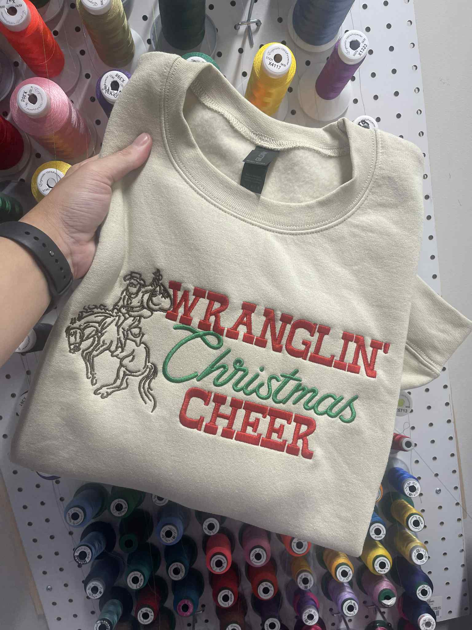 Beige sweatshirt with 'WRANGLIN' Christmas CHEER' text and graphic held against a pegboard background with spools of thread.