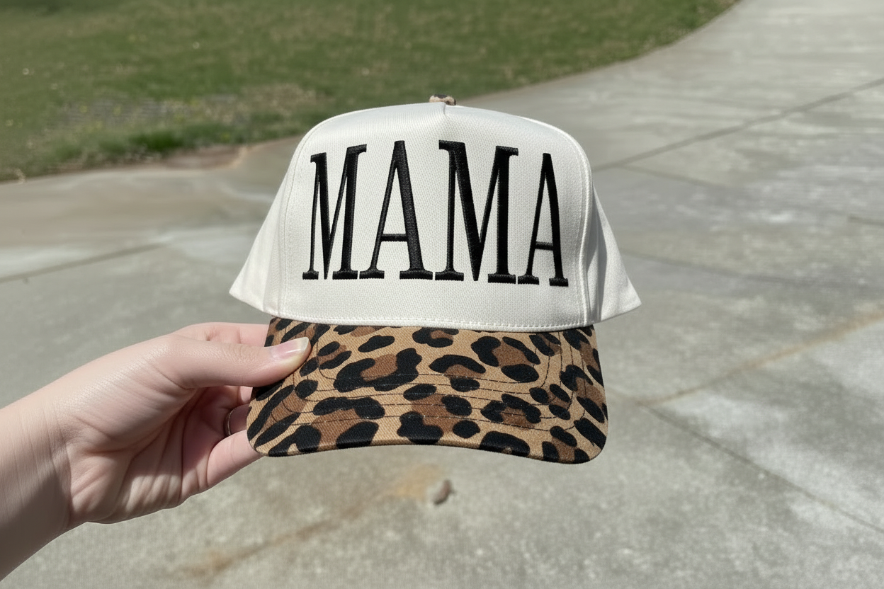 Cap with 'MAMA' text and leopard print brim held by a hand on a concrete surface.