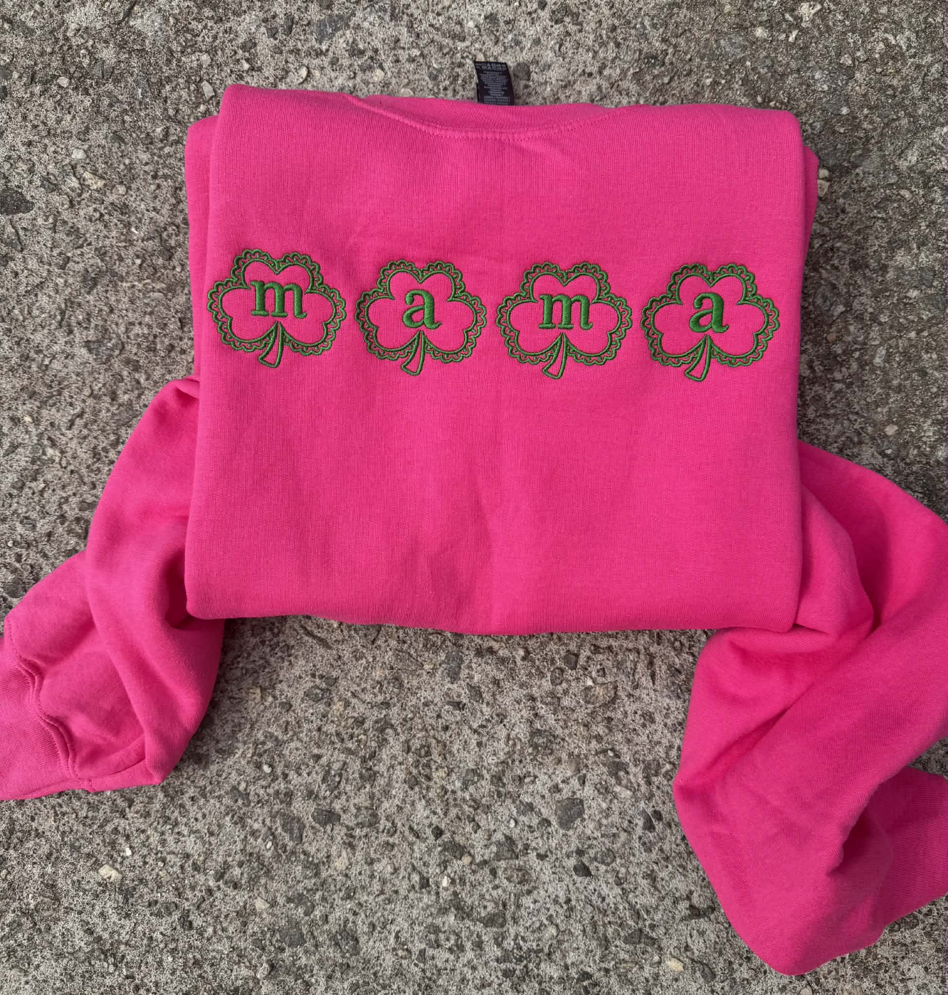 Pink sweatshirt with 'mama' printed on it, laid flat on a concrete surface.