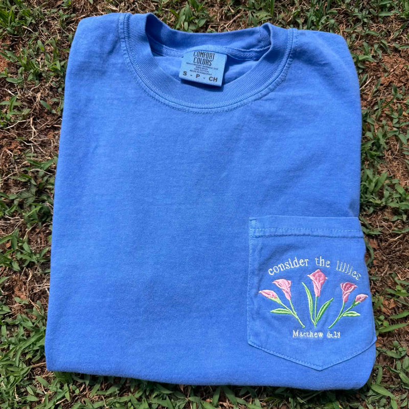 Blue Comfort Colors pocket t-shirt featuring embroidered pink lilies and the phrase "Consider the Lilies" with Matthew 6:28.