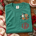 Green Comfort Colors Christmas pocket tee with embroidered milk, cookies, gingerbread, and holly above the pocket and a personalized red three-letter monogram embroidered on the pocket.
