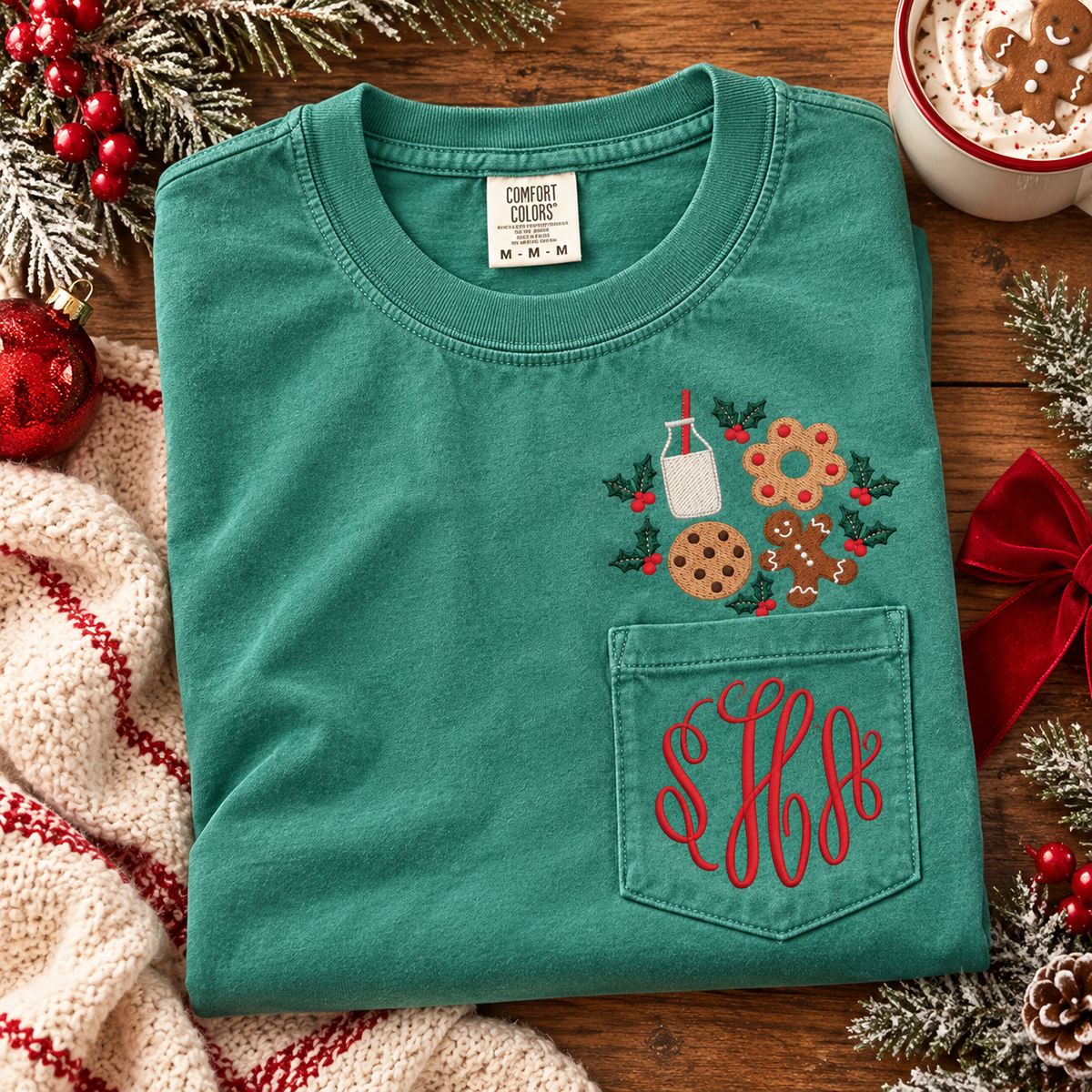 Green Comfort Colors Christmas pocket tee with embroidered milk, cookies, gingerbread, and holly above the pocket and a personalized red three-letter monogram embroidered on the pocket.