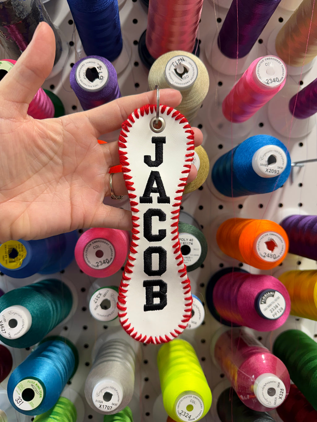 Hand holding a white and red bone-shaped object with JACOB in front of colorful spools of thread on a pegboard.
