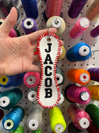 Hand holding a white and red bone-shaped object with JACOB in front of colorful spools of thread on a pegboard.
