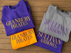 Four folded crewneck sweatshirts arranged on a wooden surface, each with large vertical team text: orange 'Granbury Pirates', purple 'Granbury Pirates' and 'Sullivan Arrows', and light gray 'Sullivan Arrows' with purple lettering.