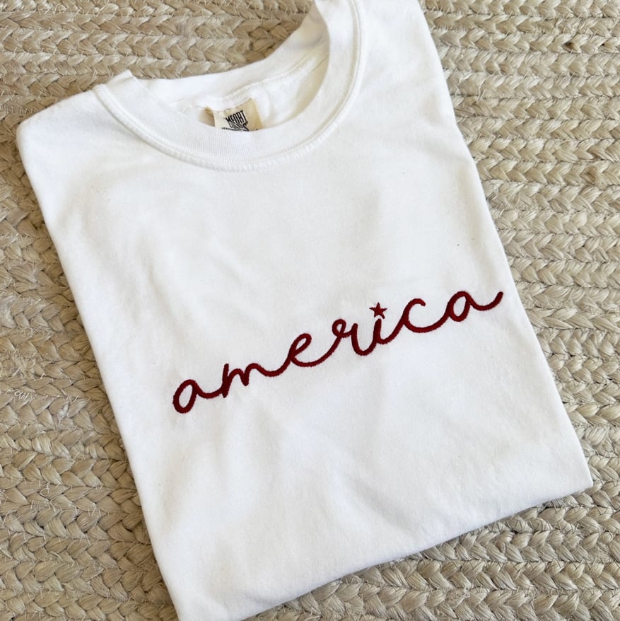 White Comfort Colors embroidered America script t-shirt featuring a patriotic star detail in red thread.