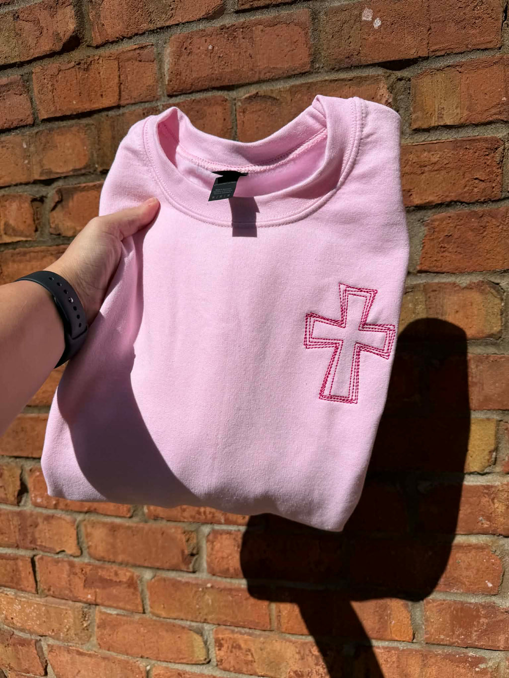 Pink sweatshirt with a cross design held in front of a brick wall