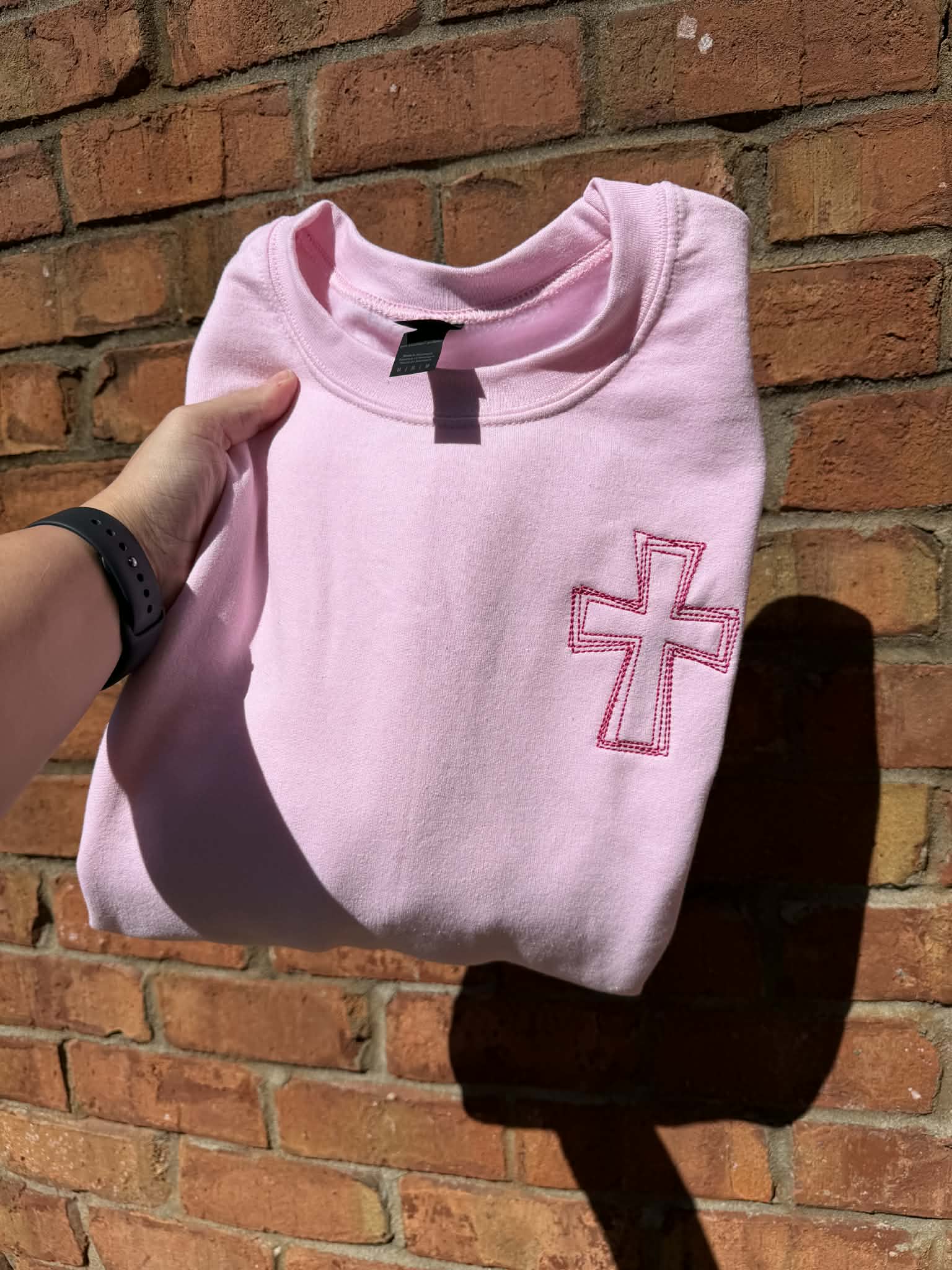 Pink sweatshirt with a cross design held in front of a brick wall