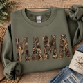 Green sweatshirt with 'MAMA' in camouflage print on a wooden surface with accessories.