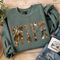 Green sweatshirt with 'WIFE' in camouflage print, placed on a wooden surface with jeans and a mug.