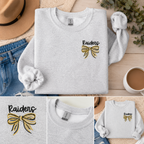 Gray sweatshirt with 'Raiders' and a bow design, placed on a wooden surface with jeans and a coffee cup.