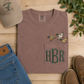 Brown shirt with duck design and 'HBR' initials, surrounded by jeans, shoes, and a cap on a wooden surface.
