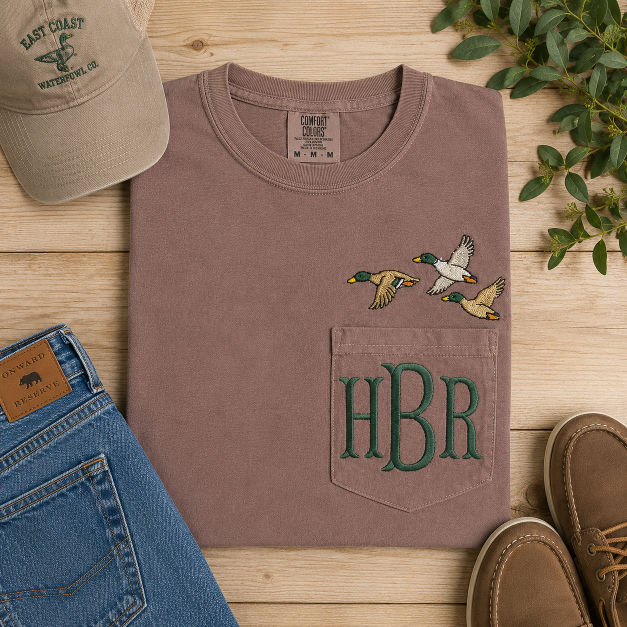 Brown shirt with duck design and 'HBR' initials, surrounded by jeans, shoes, and a cap on a wooden surface.