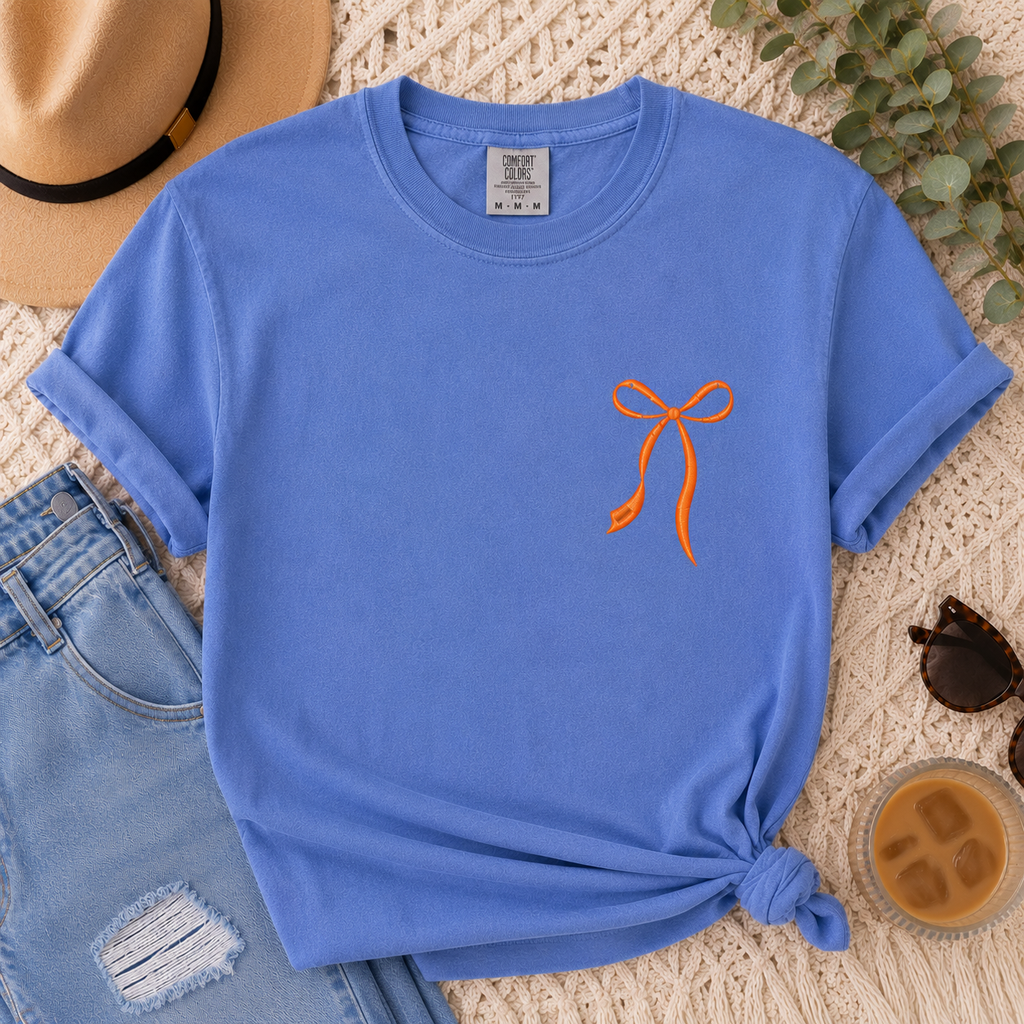 Blue t-shirt with orange bow design on a textured surface with jeans, hat, and coffee.