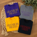 Four folded crewneck sweatshirts arranged on a woven round mat over a wooden surface. The sweatshirts are in purple, gray, yellow, and black, each printed with team-style text: 'GRANBURY PIRATES' in gold, 'SULLIVAN ARROWS' in purple, 'GRANBURY PIRATES' in dark blue, and 'CHEROKEE CHIEFS' in red.
