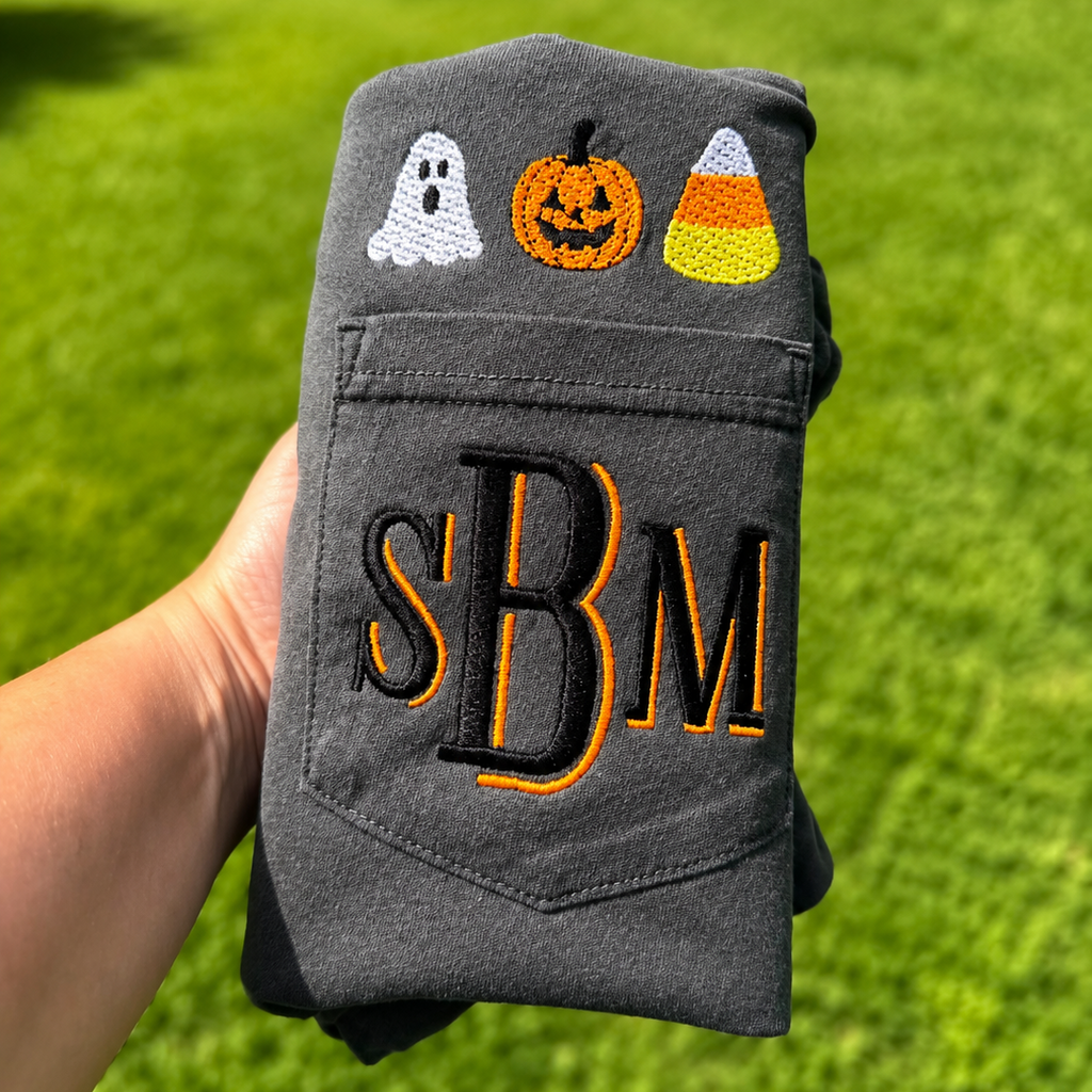 A folded dark gray sweatshirt held by a hand against a green grass background. The sweatshirt features embroidered Halloween motifs, including a white ghost, an orange jack-o'-lantern, and a candy corn, with large black and orange embroidered letters 'SBM' on the pocket.