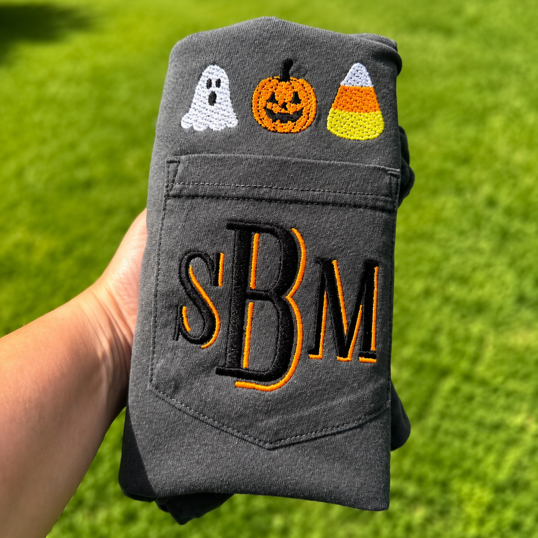 A folded dark gray sweatshirt held by a hand against a green grass background. The sweatshirt features embroidered Halloween motifs, including a white ghost, an orange jack-o'-lantern, and a candy corn, with large black and orange embroidered letters 'SBM' on the pocket.