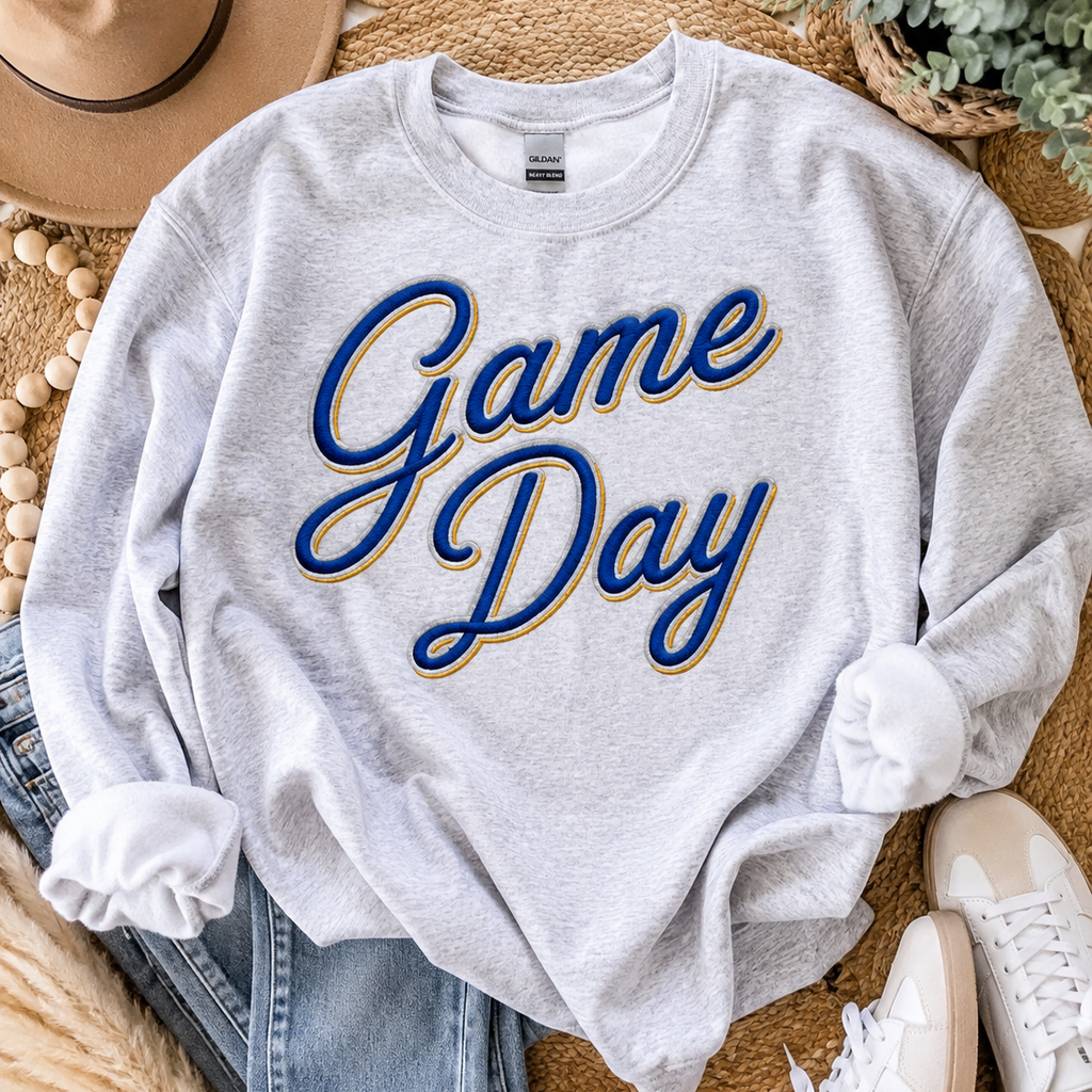 A light heather gray crewneck sweatshirt with 'Game Day' printed in blue cursive letters with a gold outline, styled in a flat lay with jeans, white sneakers, a hat, and a plant on a woven rug.