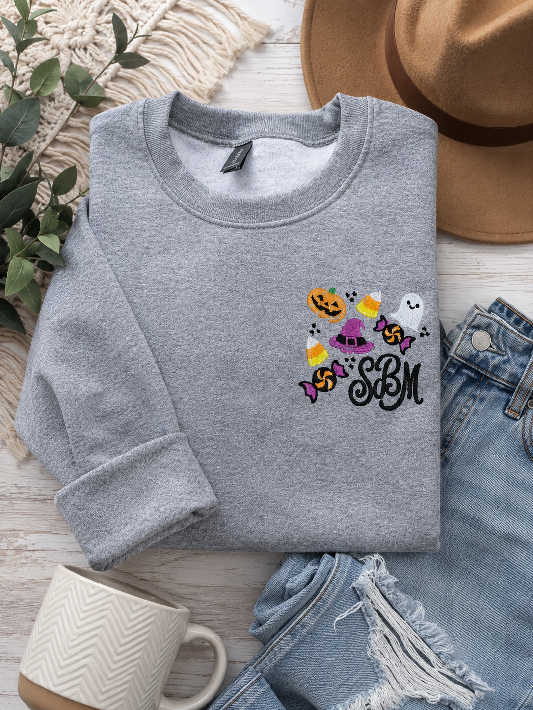Gray embroidered Halloween sweatshirt featuring a personalized black monogram surrounded by a pumpkin, ghost, witch hat, candy corn, and Halloween candy embroidery design on the left chest.