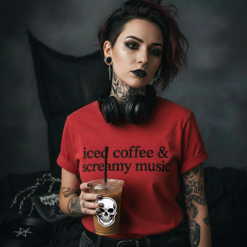 A person with short dark hair, tattoos, and black eye makeup wears a red graphic T-shirt with the text 'iced coffee & creamy music' and holds an iced coffee cup with a skull sticker. Black headphones hang around the neck, and a dark gothic-style setting with a draped cloth is in the background.