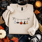 A beige crewneck sweatshirt with a Halloween-themed graphic of hanging ghosts, bats, a pumpkin, a witch hat, and candy corn, styled in a flat lay with autumn props and a 'Stay Spooky' sign.