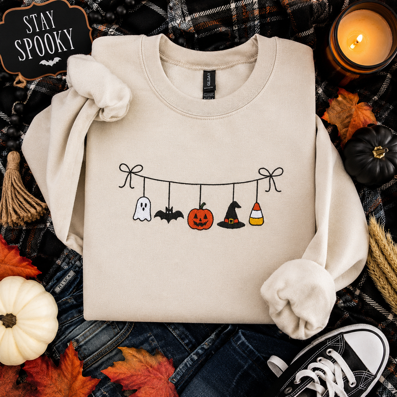 A beige crewneck sweatshirt with a Halloween-themed graphic of hanging ghosts, bats, a pumpkin, a witch hat, and candy corn, styled in a flat lay with autumn props and a 'Stay Spooky' sign.