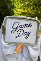 A light gray sweatshirt held up outdoors by a hand, with a large embroidery hoop covering most of the front. The hoop shows the words “Game Day” in blue script with a gold outline.