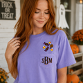 A woman with long red hair wearing a lavender purple short-sleeve t-shirt with a pocket featuring embroidered candy corn, a witch's hat, and the initials 'sBM'. The background shows a cozy, decorated Halloween porch with pumpkins, a ghost decoration, and a 'Trick or Treat' sign.