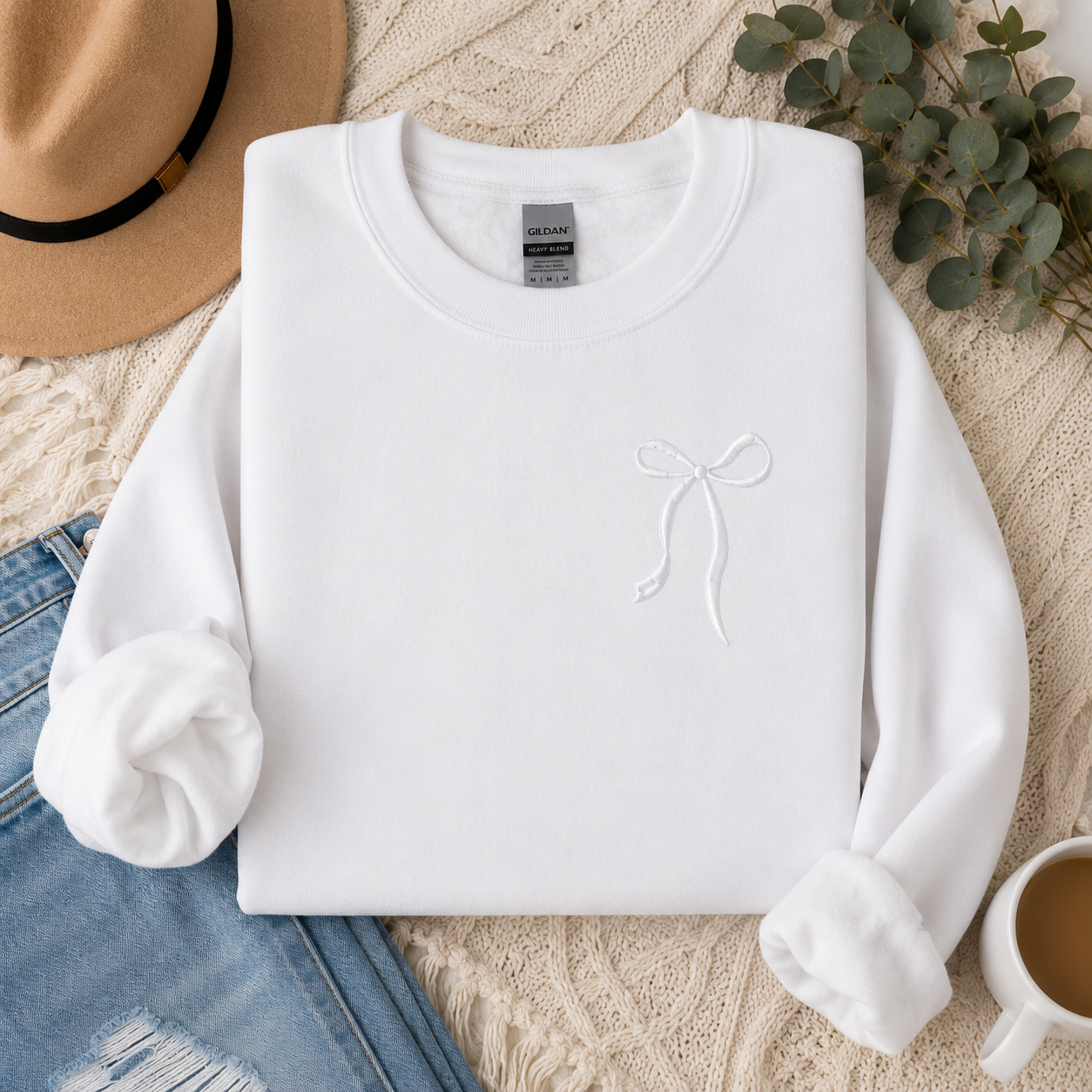 White sweatshirt with a logo on a textured surface with a hat, jeans, and coffee cup.