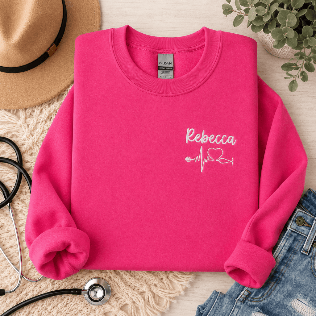Pink sweatshirt with 'Rebecca' design on a textured surface with a hat, jeans, and stethoscope.