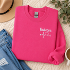 Pink sweatshirt with 'Rebecca' and heart stethoscope design on a textured surface with jeans and a hat.