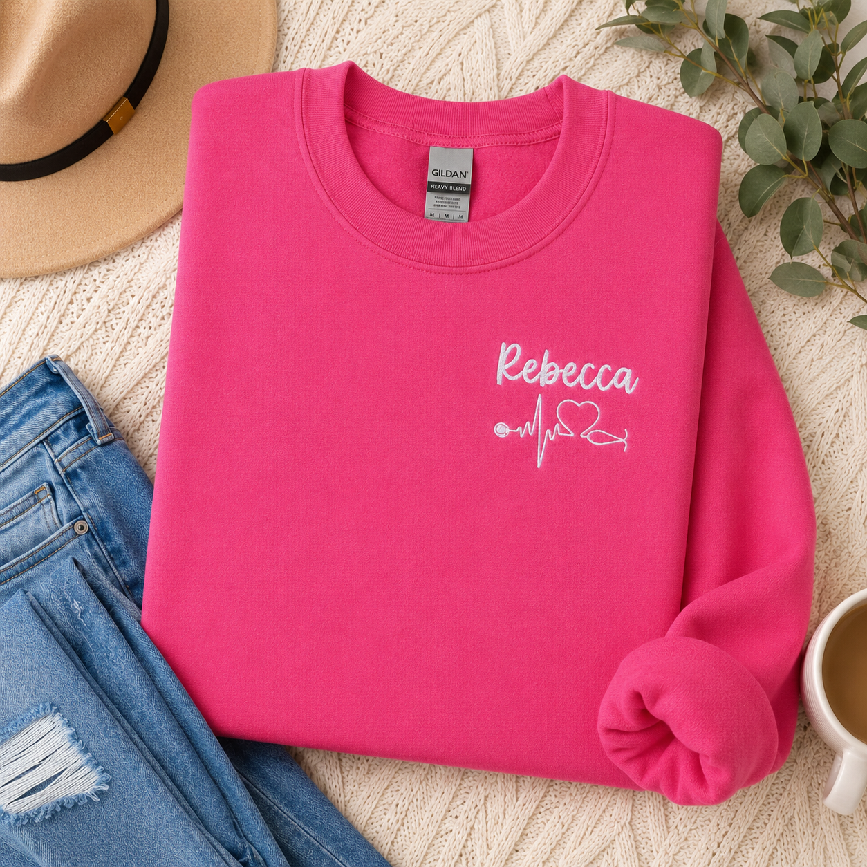 Pink sweatshirt with 'Rebecca' and heart stethoscope design on a textured surface with jeans and a hat.