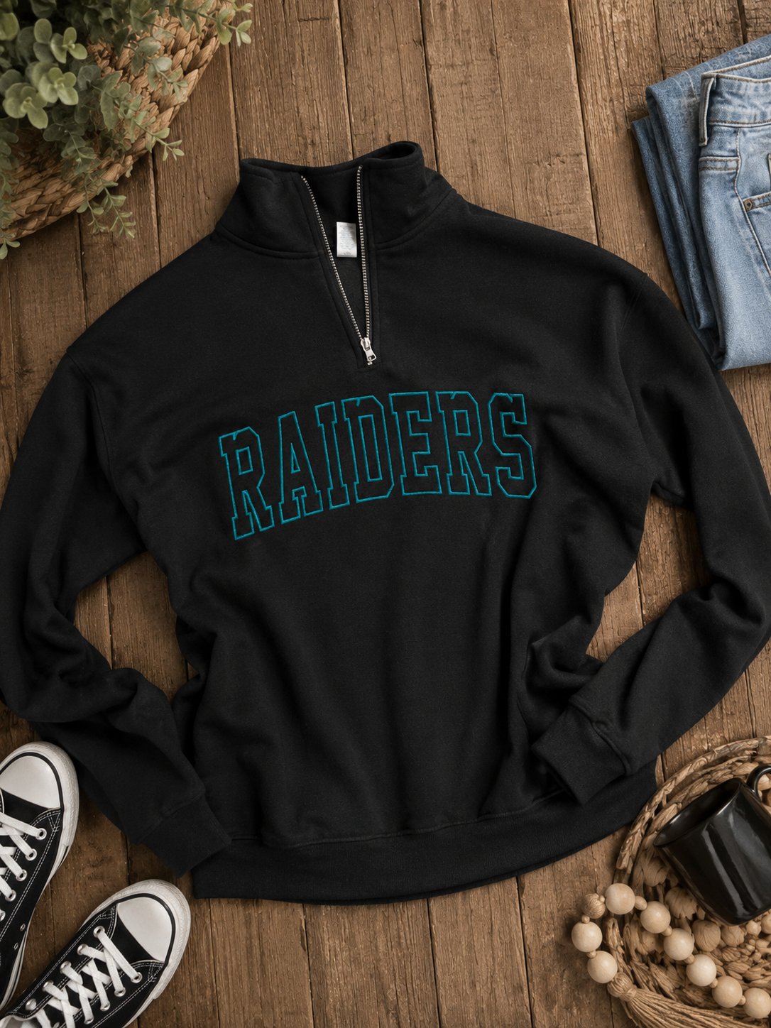 A black long-sleeve quarter-zip sweatshirt with 'RAIDERS' printed across the chest in teal outlined block letters, styled in a flat lay on a wooden surface with jeans, sneakers, and decorative woven accents.