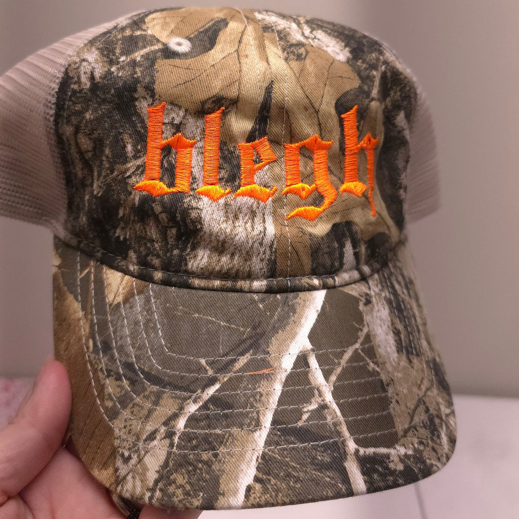 Camouflage-patterned cap with orange text held by a hand against a neutral background