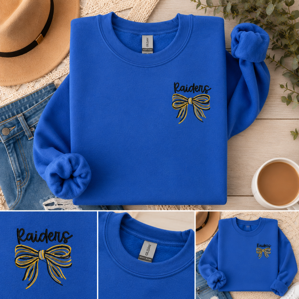 Blue sweatshirt with 'Raiders' and bow design, placed on a wooden surface with a hat, jeans, and coffee cup.