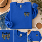 Blue sweatshirt with 'Raiders' and bow design, placed on a wooden surface with a hat, jeans, and coffee cup.
