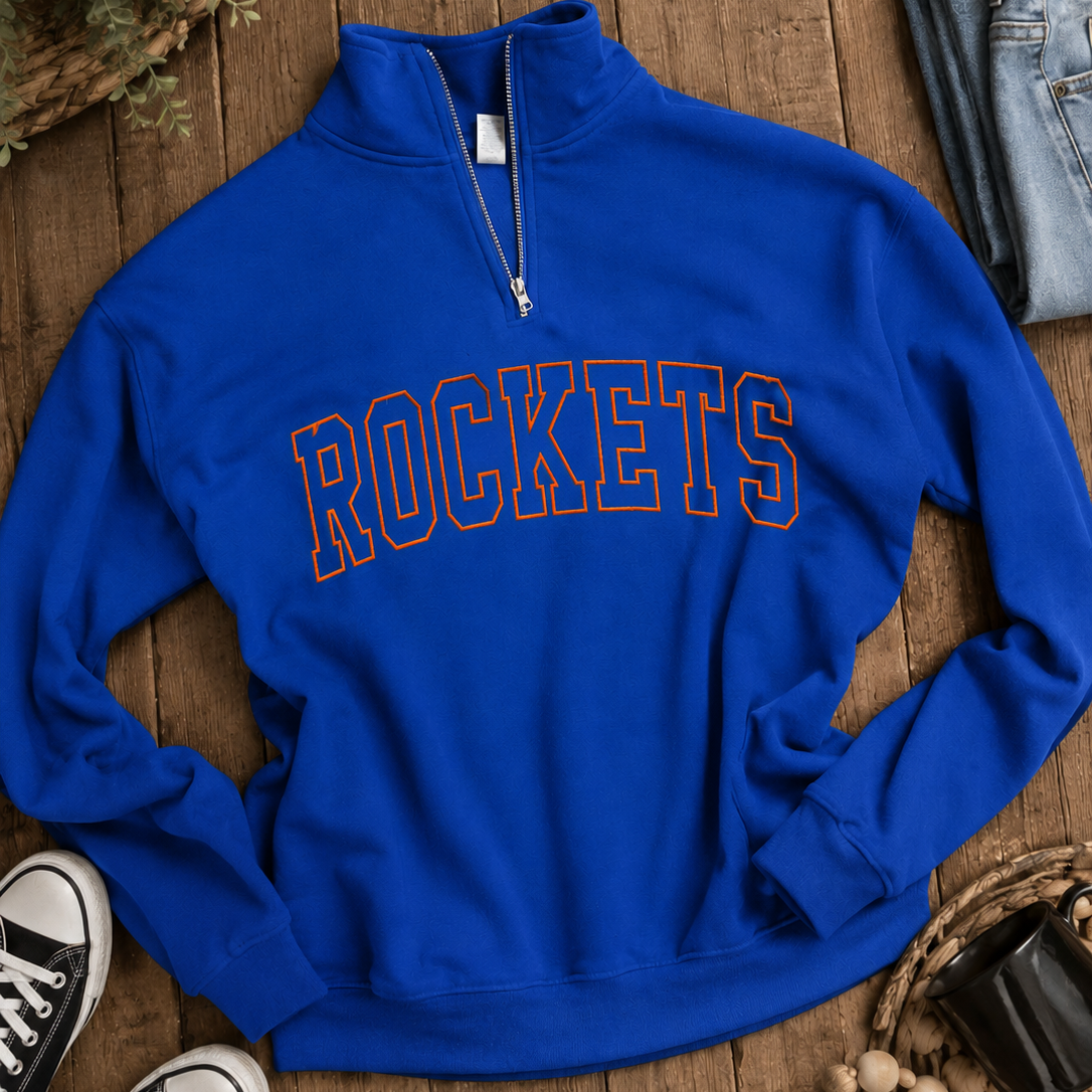 A royal blue quarter-zip sweatshirt with 'ROCKETS' printed across the chest in an arched varsity font with an orange outline. The sweatshirt is styled in a flat-lay arrangement on a rustic wooden background, surrounded by folded blue jeans, black sneakers, and decorative woven baskets.