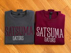Two folded short-sleeve T-shirts laid flat on a wood floor: one burgundy with large white 'SATSUMA GATORS' text, and one dark gray with large red 'SATSUMA GATORS' text.