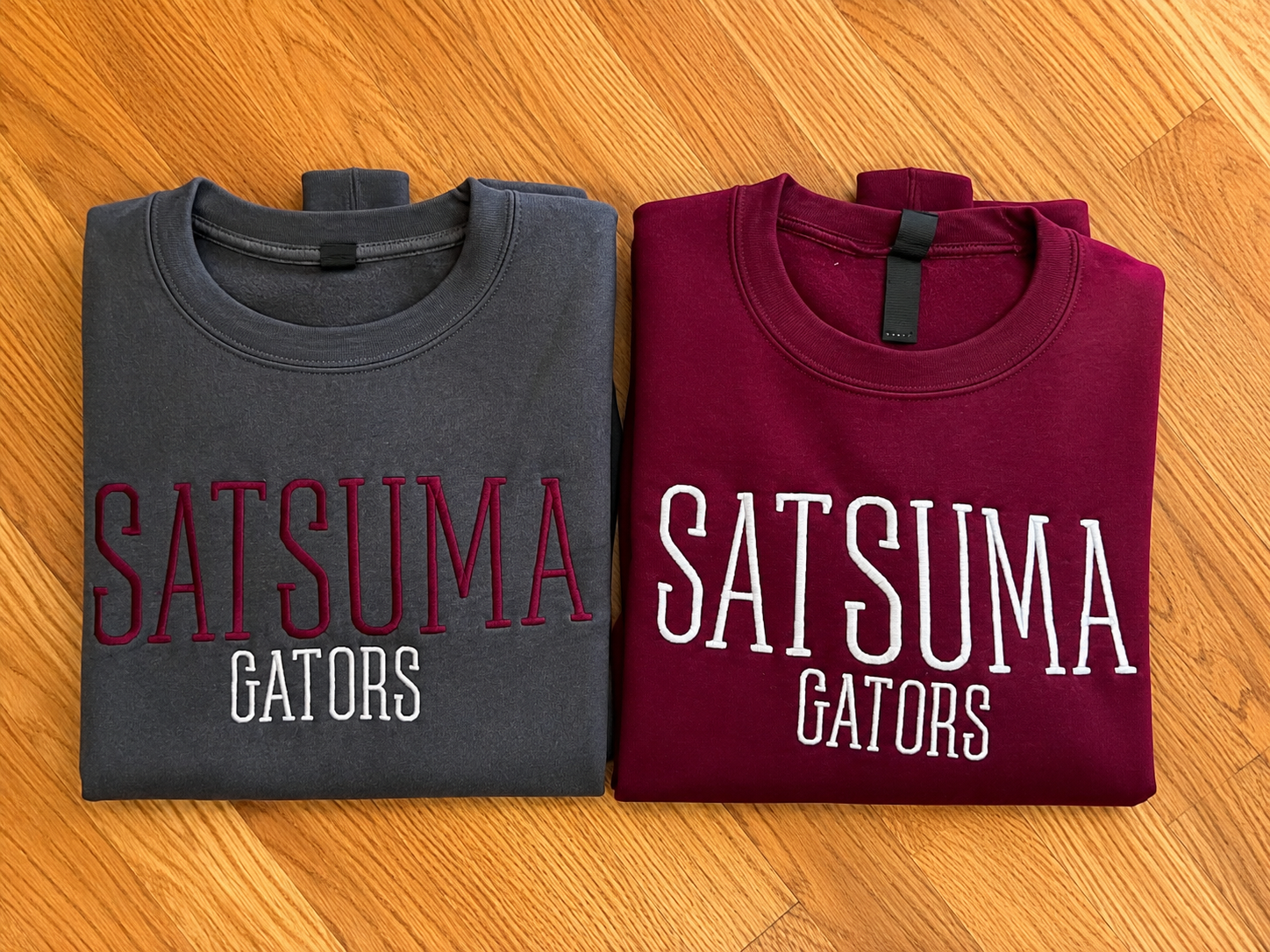 Two folded short-sleeve T-shirts laid flat on a wood floor: one burgundy with large white 'SATSUMA GATORS' text, and one dark gray with large red 'SATSUMA GATORS' text.