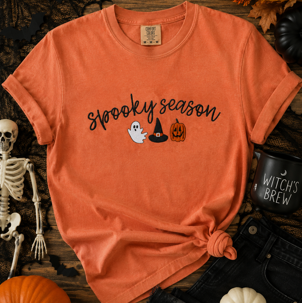 Orange t-shirt with 'Spooky Season' text and Halloween graphics on a wooden surface with Halloween decorations.