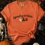 Orange t-shirt with 'Spooky Season' text and Halloween graphics on a wooden surface with Halloween decorations.