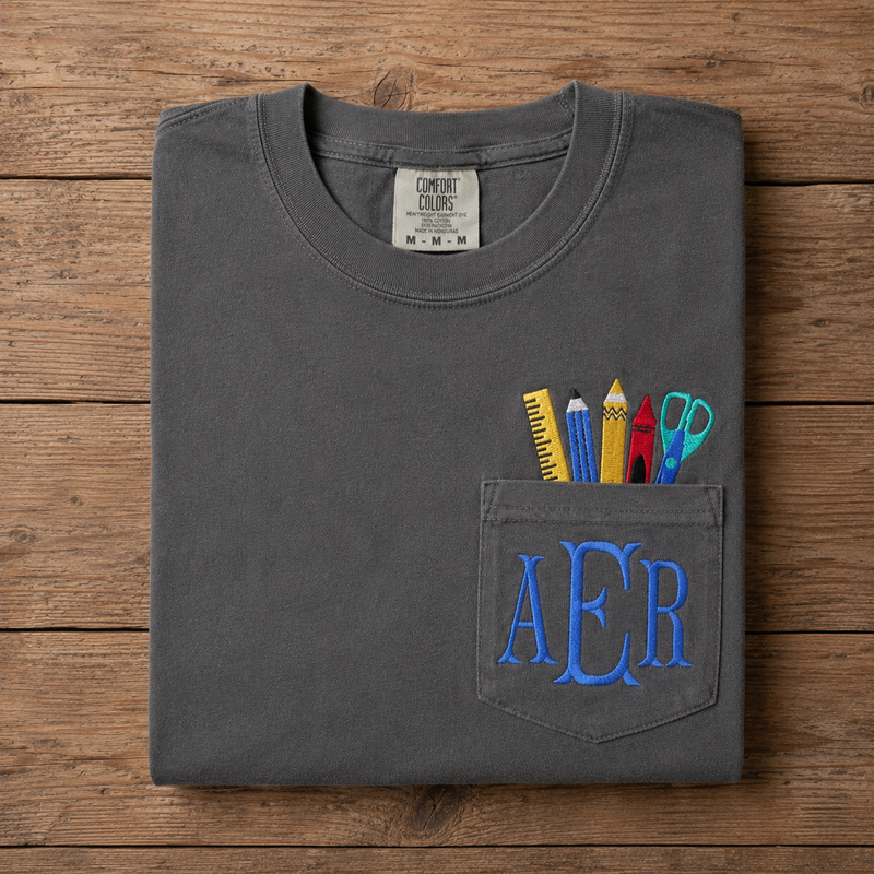 Folded gray t-shirt with pencil case design and 'AER' fishtail monogram on a wooden surface