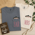 Two folded shirts with fish designs and initials, next to a wooden sign about lake life, on a wooden surface.